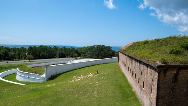 A canon peers over a brick wall with the coastline in the distance. — Gulf Islands National Seashore, NPS